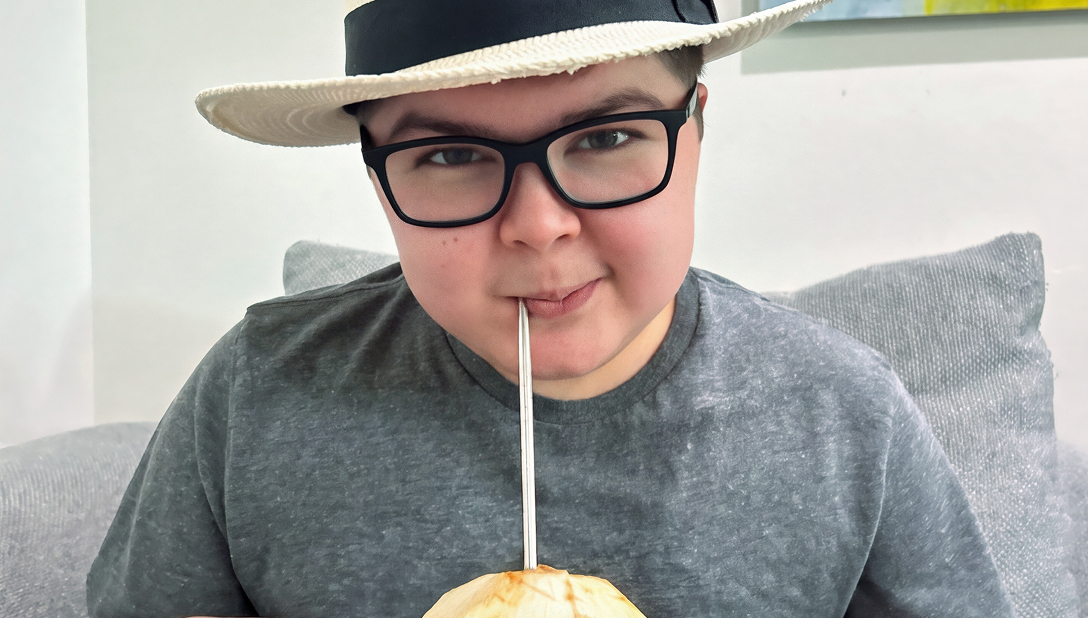 Boy wearing glasses and a light-colored fedora hat sitting on a couch indoors, drinking from a green coconut through a straw. Text reads in the bottom right corner "Alex, Age 19 — Living with Duchenne."