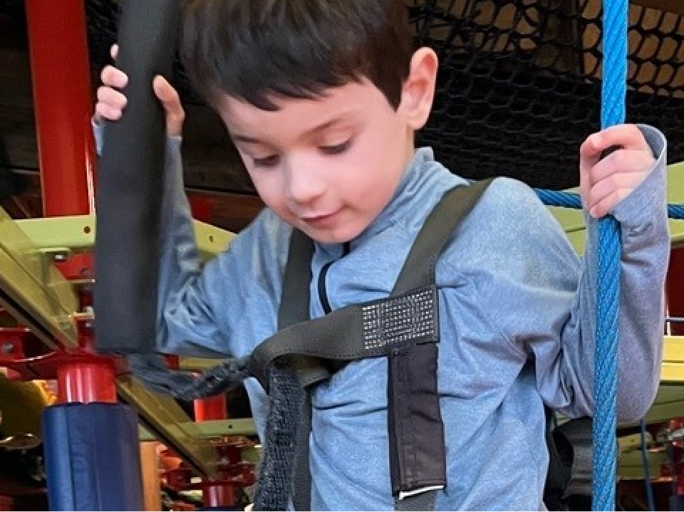 Boy wearing a safety harness navigating a suspended rope obstacle course indoors, stepping across wooden planks while holding onto blue support ropes. Small text in the bottom left corner reads "Tripp, Age 6— Living with Duchenne"